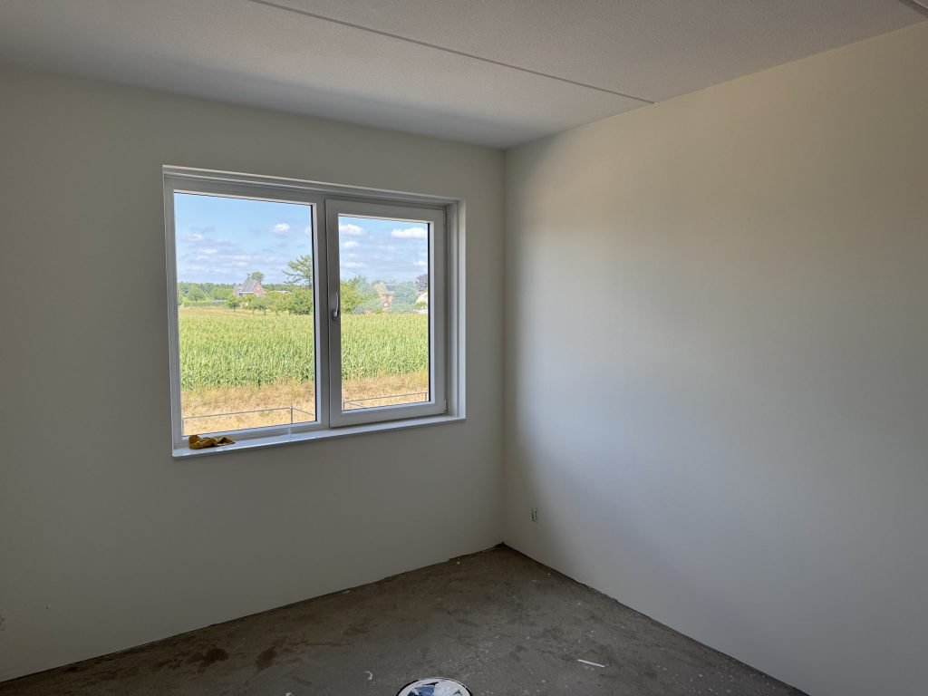 Lege nieuwbouwkamer met gladde lichte wanden en een raam dat uitkijkt op een maisveld